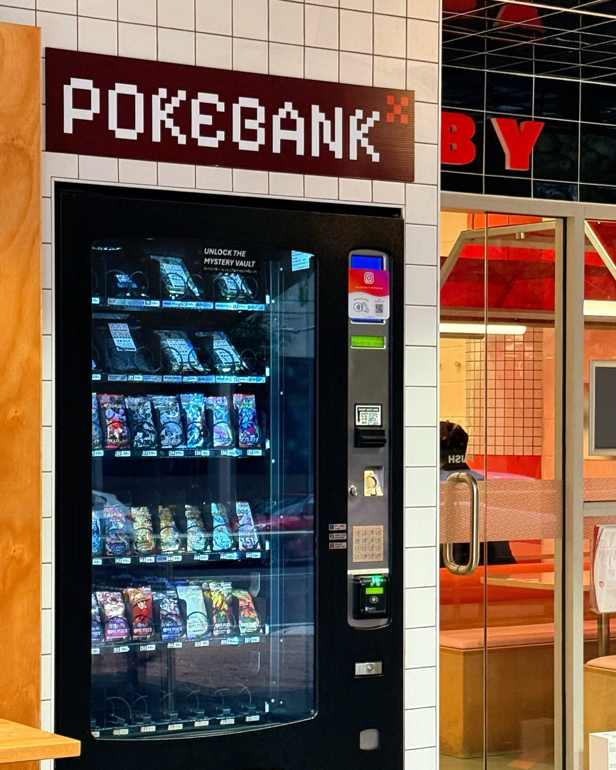 A black automatic vending machine labeled 'POKEGANK' stands against a white tiled wall next to a glass entrance. The vending machine is stocked with packaged snack items and has a coin and card reader on the right side.